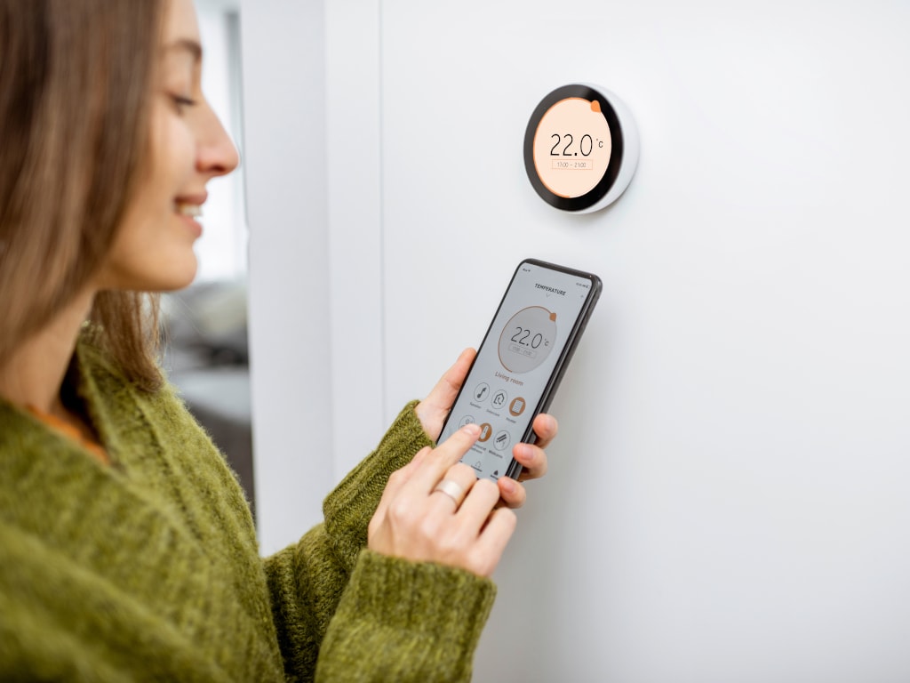 Junge Frau von der Seite bedient Smartphone und steht vor weißer Wand mit rundem Thermostat