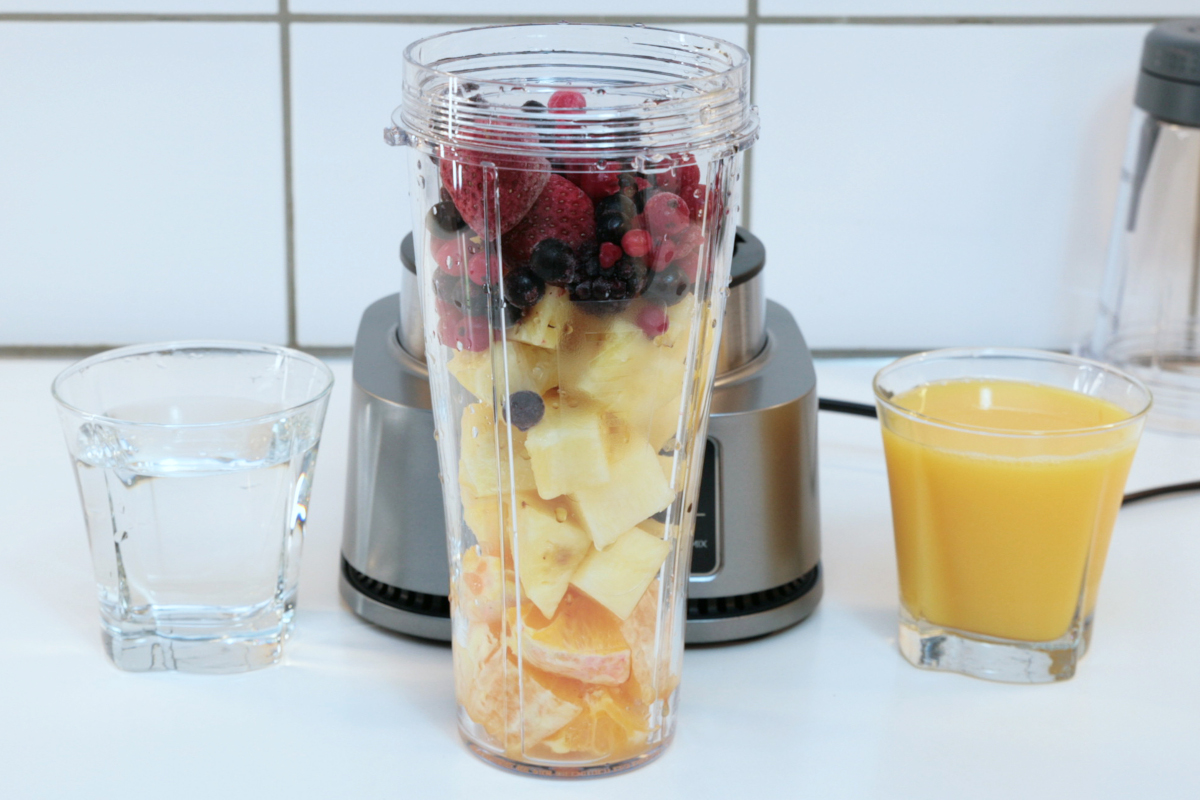 Mixer Becher mit Ananas, Beeren und Orange gefüllt vor Standmixer, daneben kleines Glas mit orangenem Smoothie
