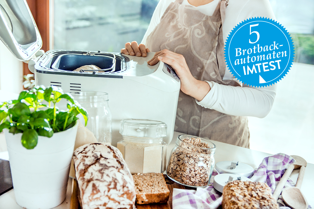 Frau ab Hals abwärts im Bild hält Hände an weißem geöffneten Brotbackautomaten, der auf Tisch mit Brot und Zutaten steht