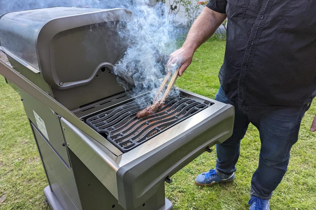 Mann steht neben silbernem Gasgrill und wendet qualmende Wurst