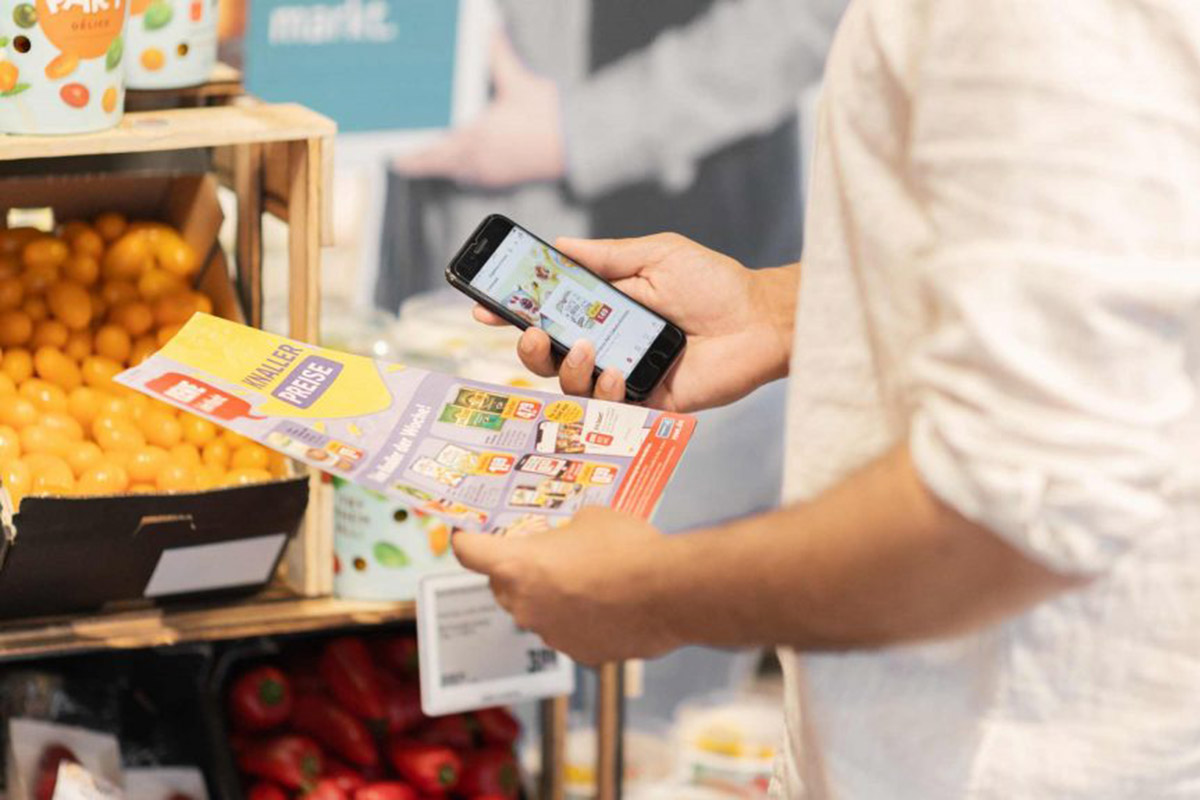 Eine Person hält einen Werbeprospekt von Rewe in der Hand sowie die Rewe App auf dem Handy.