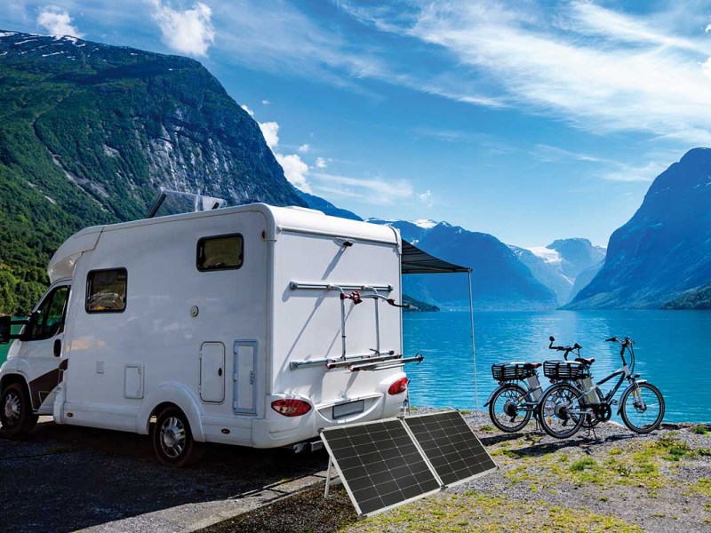 Ein weißes Wohnmobil, zwei Elektofahrräder und die neuen revolt Solarpaneele stehen vor einer Berglandschaft mit großem See.