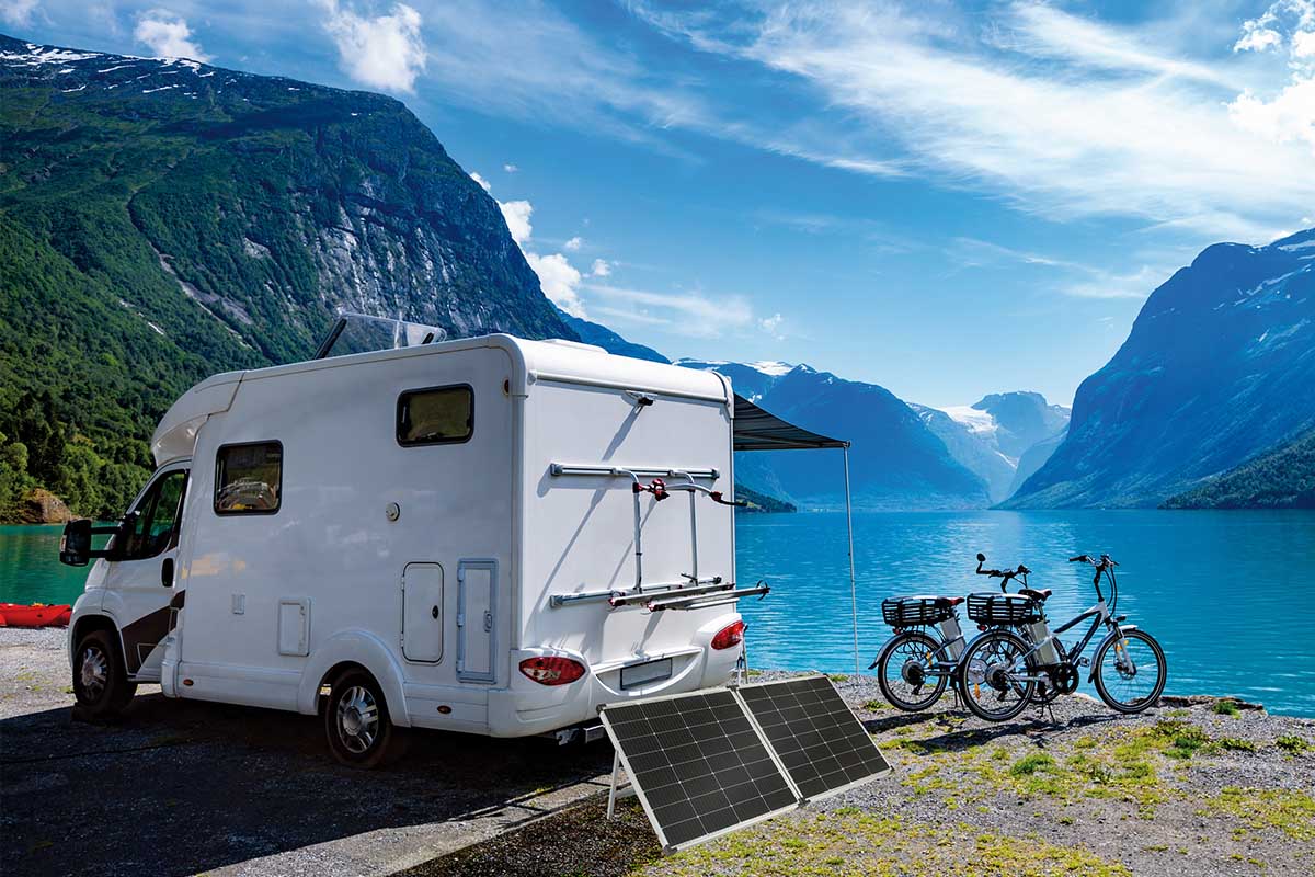 Ein weißes Wohnmobil, zwei Elektofahrräder und die neuen revolt Solarpaneele stehen vor einer Berglandschaft mit großem See.