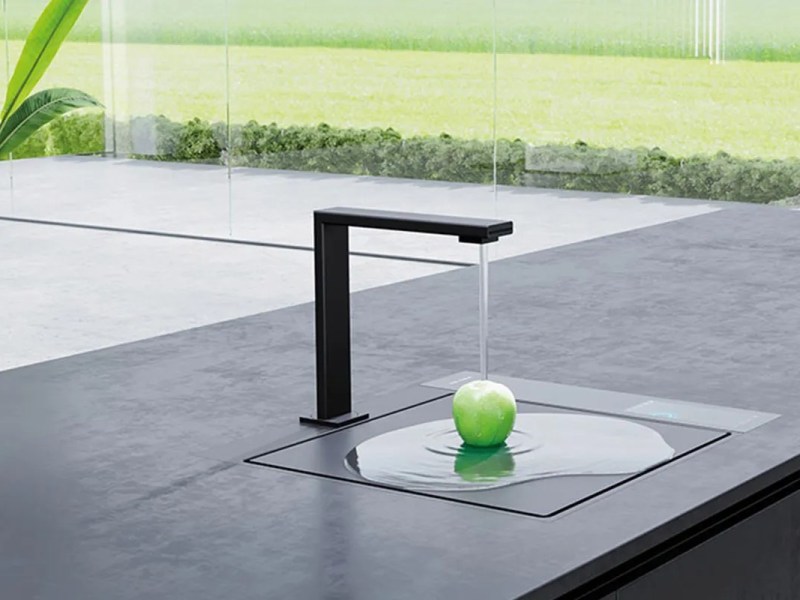 Es ist die neue Solitaire Waterbase Spüle mit abgedecktem Spülbecken zu sehen. Ein Apfel liegt quasi auf der Arbeitsplatte und wird vom Wasser aus dem Hahn abgespült.