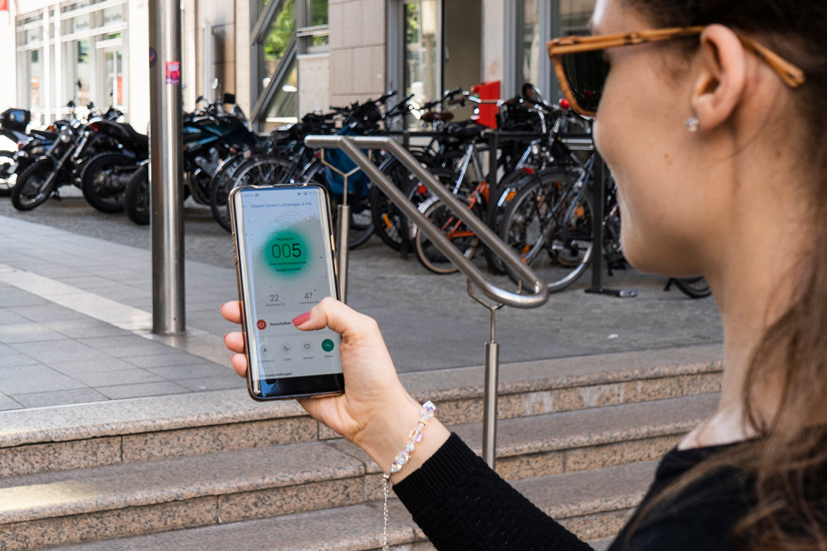 Eine Person mit Sonnenbrille schaut auf ein Smartphone mit Luftfilter-App. Drumherum Straßenumgebung.