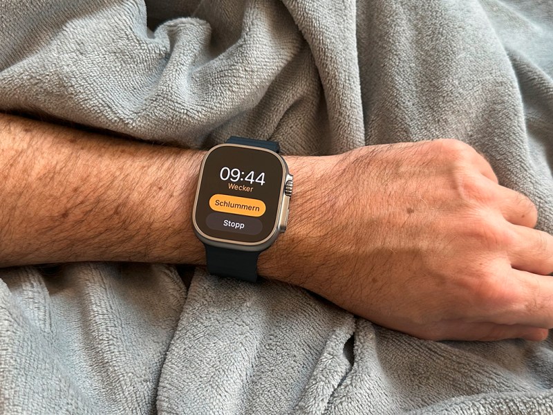 Männerarm liegt auf grauer Decke mit dunkler Smartwatch an Gelenk