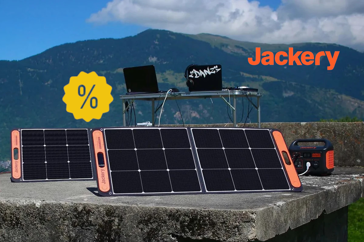 Aufgestellte Solarpanel vor Powerstation und Tisch mit Computer auf steinplatte vor Bergkulisse mit gelben Prozentzeichen links und Jackery Logo rechts