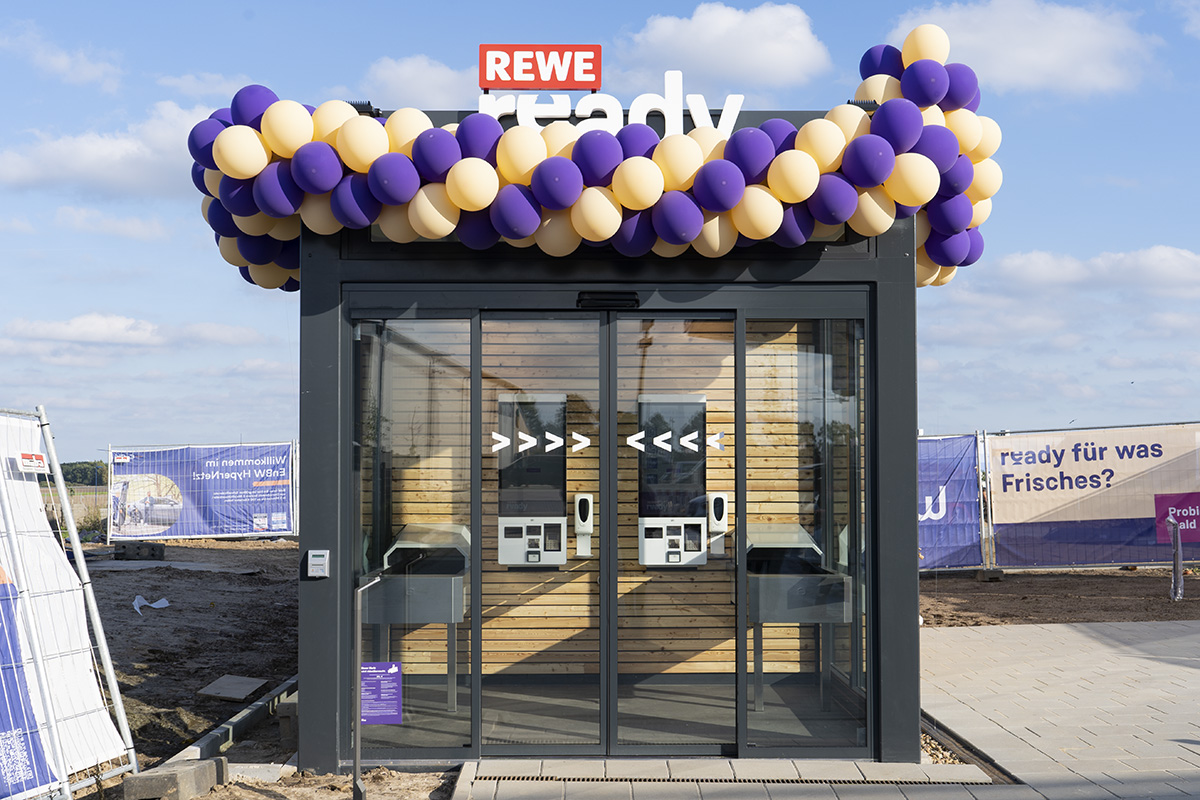 Rewe ready besteht aus einem holzverkleideten Lager und einem kleinen, verglasten Eingangsbereich mit je zwei Tochscreens und Laufbändern. Zur Eröffnung war die Mini-Filiale mit gelben und lilafarbenen Luftballons dekoriert.