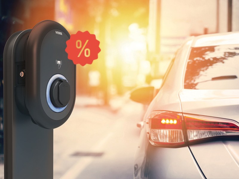 Links schwarze stehende Vestel-Wallbox mit rotem Prozentzeichen vor Straße in goldenem Sonnenlicht, rechts weißes Auto von hinten