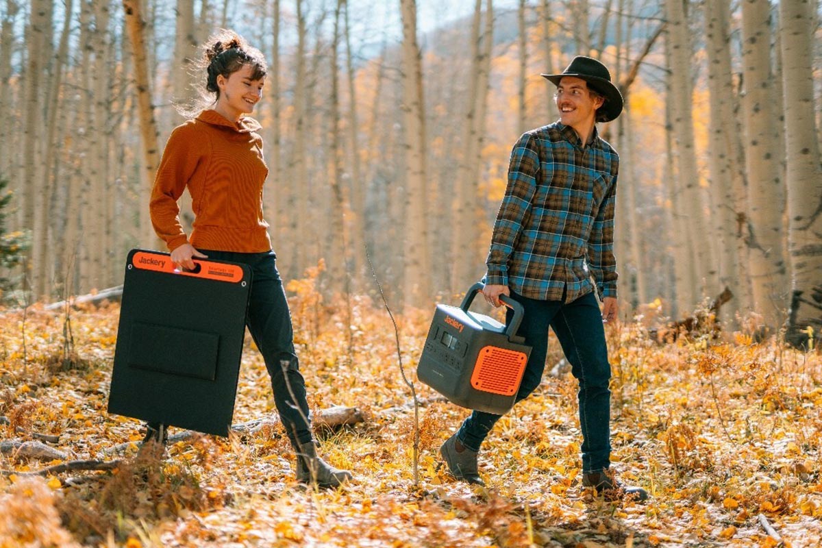 Mann und Frau gehen durch Wald voller Laub mit Solar Panel und Power Station in der Hand