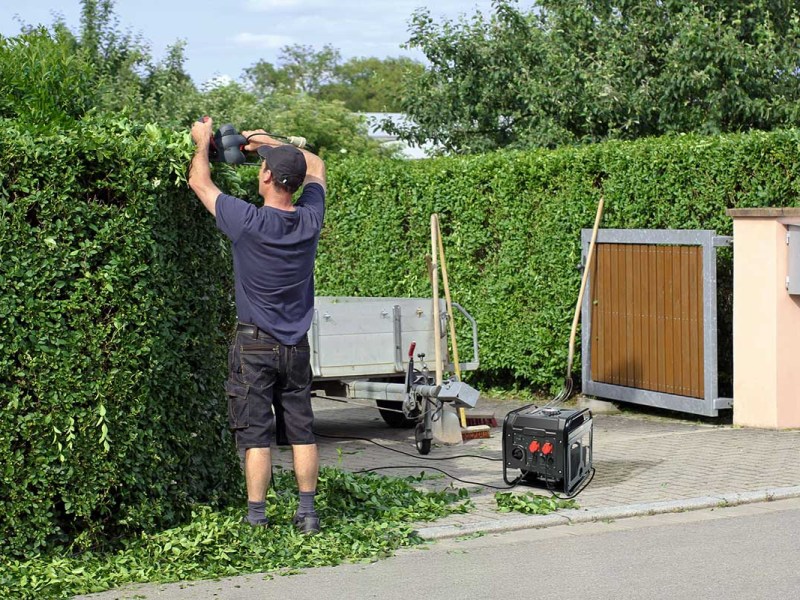Ein Mann schneidet eine Hecke. Als Stromquelle dient der Revolt BIG-275 Benzingenerator.
