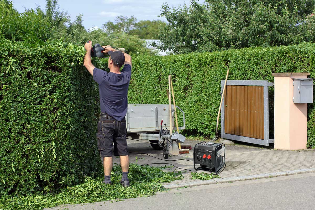 Ein Mann schneidet eine Hecke. Als Stromquelle dient der Revolt BIG-275 Benzingenerator.