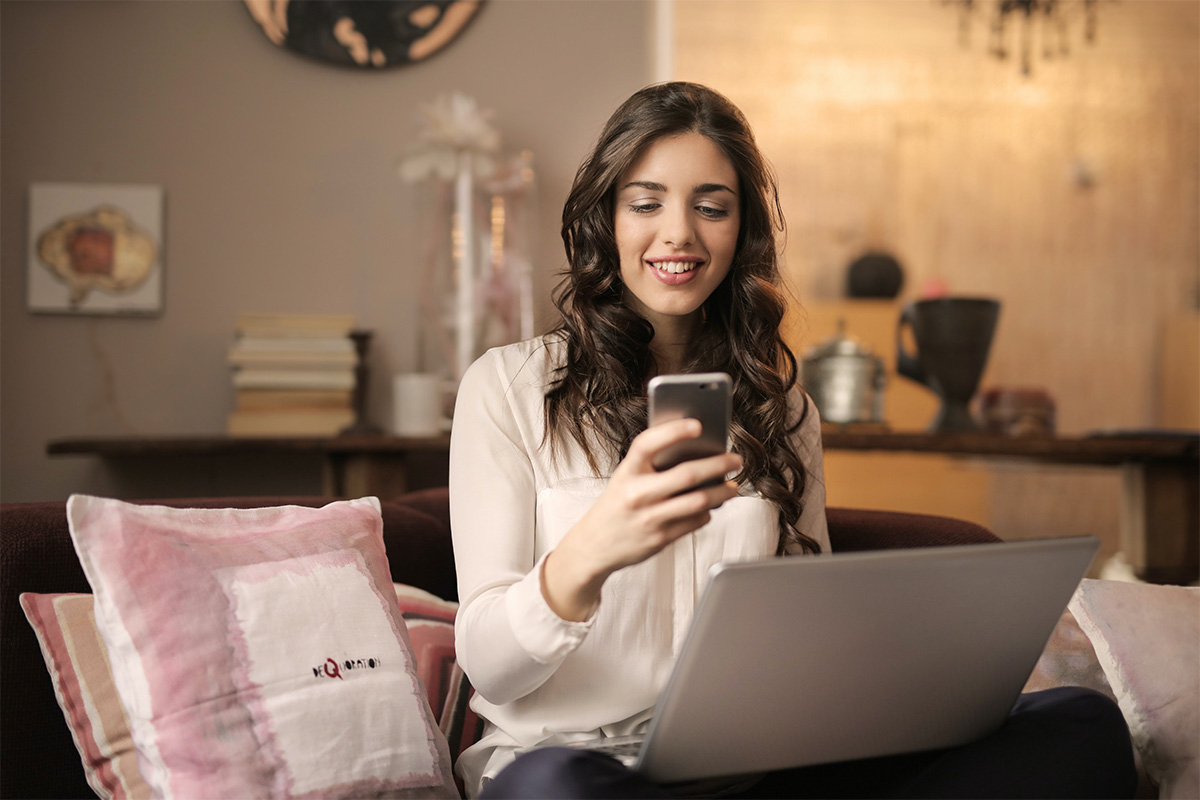 Eine Frau hat einen Laptop auf dem Schoß und schaut lächelnd auf ein Smartphone.