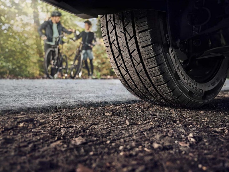 Autoreifen auf einer Schotterpiste von unten im Anschnitt, im hintergrund sieht man zwei Kinder auf einem Fahrrad