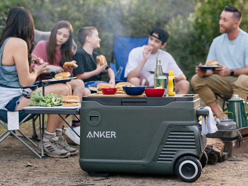 Mehrere Personen an einem Camping-Esstisch im Freien. Im Vordergrund die Anker EverFrost Kühlbox.