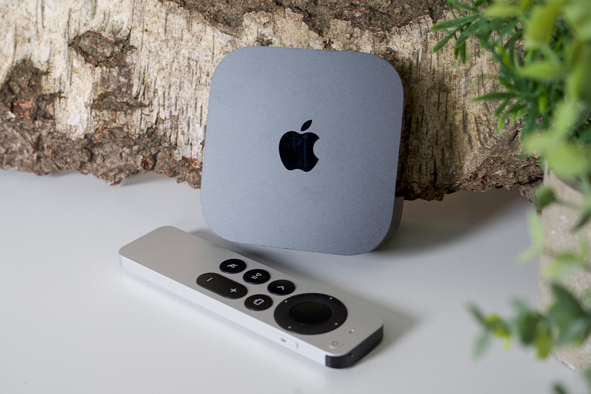 Apple TV 4K mit Fernbedienung vor einem Holzstück auf weißem Untergrund.