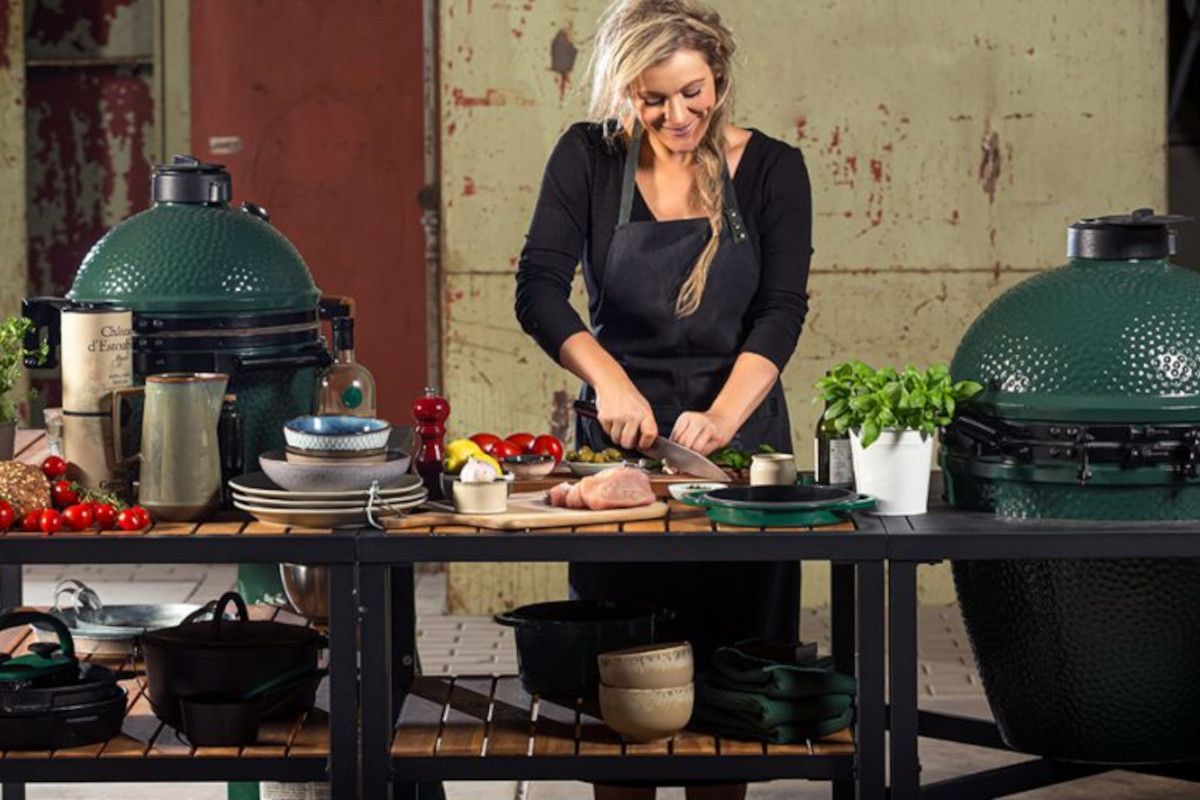 Blonde Frau mit schwarzer Schürze an Kochtisch bereitet Lebensmittel zu. Links und rechts zwei große ovale grüne Keramikgrills