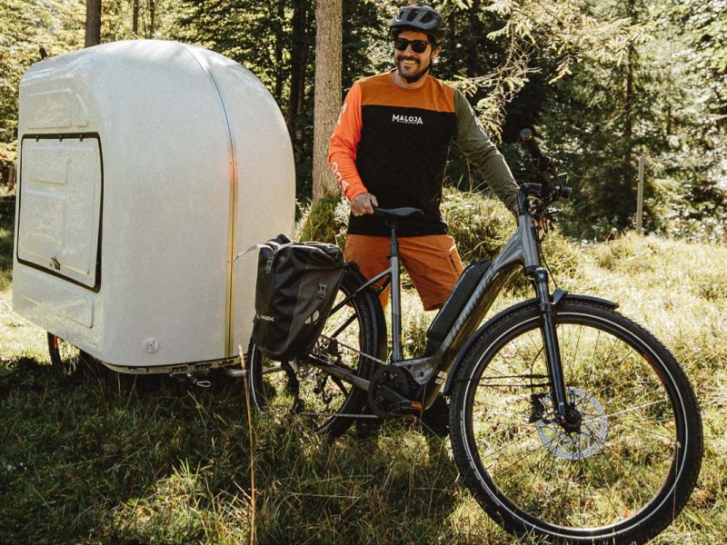 Mann steht neben seinem E-Bike im Wald, hinter seinem Rad ist ein kleiner Wohnmobil-Anhänger, der vom Rad gezogen wird