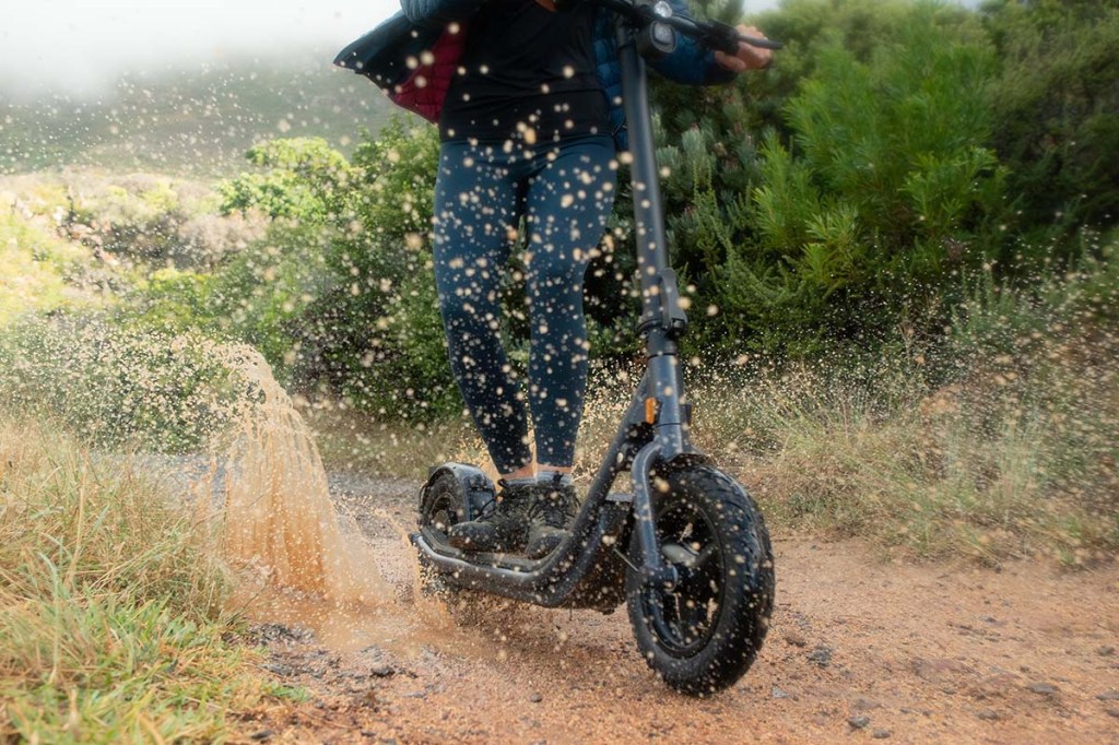 Person im Anschnitt, fährt mit E-Scooter durch eine Fütze