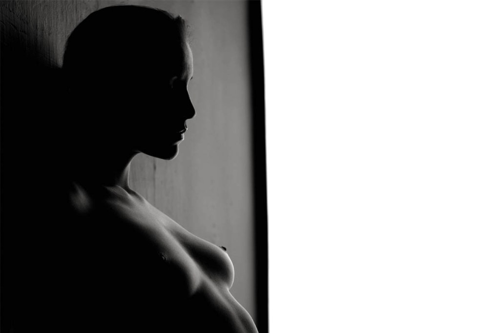 Schwarz-weiß-Aufnahme einer Frau mit nacktem Oberkörper, die an einer Wand lehnt.