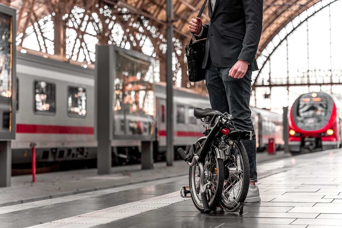 Man steht mit Faltrad auf Bahnsteig. Dahinter einfahrende Züge.