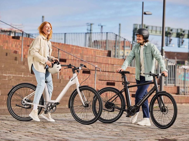 Zwei Menschen stehen neben ihren E-bikes lachend auf einem Platz.