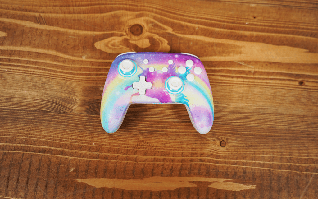 Ein sehr bunter Controller liegt auf einem Holztisch. Das Gerät hat auf der Oberseite ein Regenbogen-Motiv sowie eine Einhorn-Silhouette. 