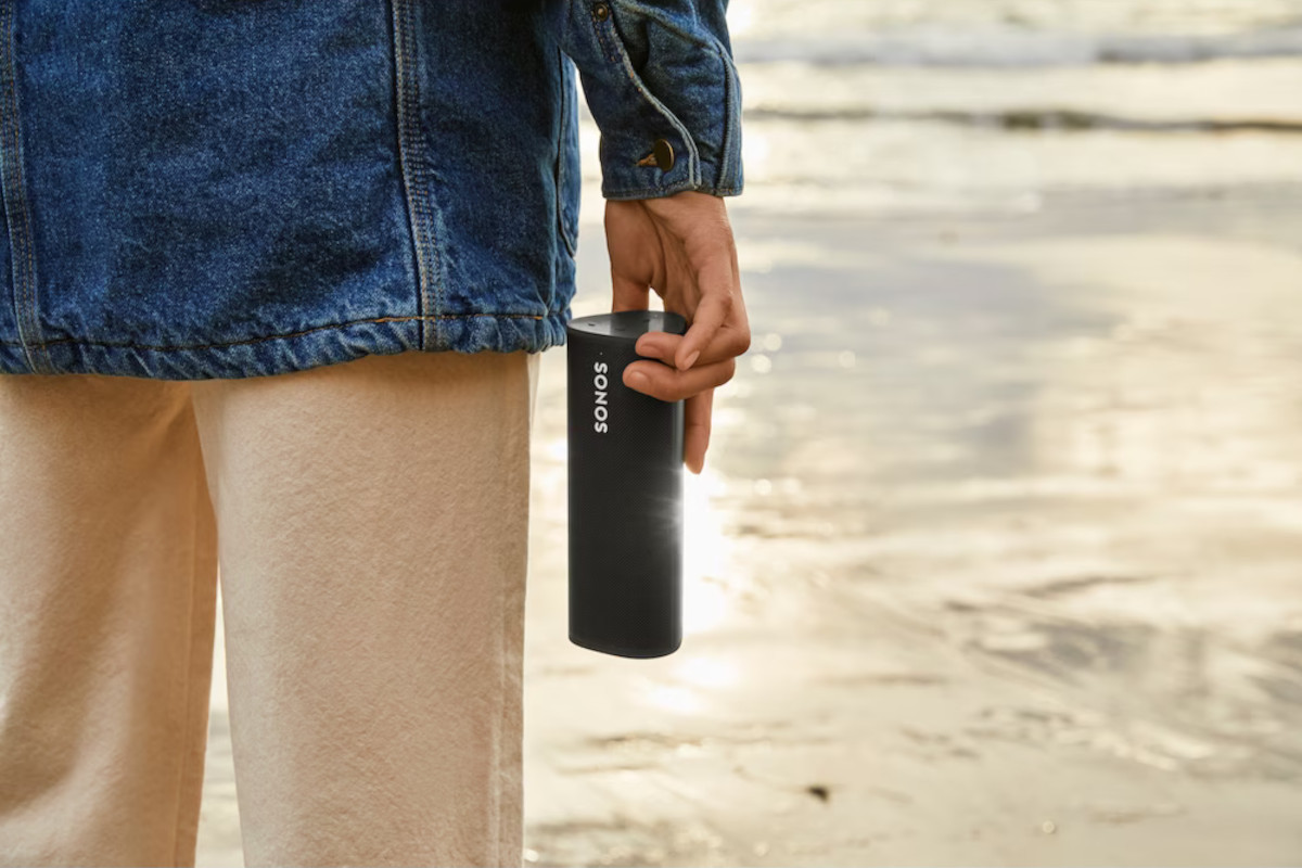 Person in beiger Hose und Jeansjacke hält schwarzen Sonos-Speaker in der Hand und steht am Strand