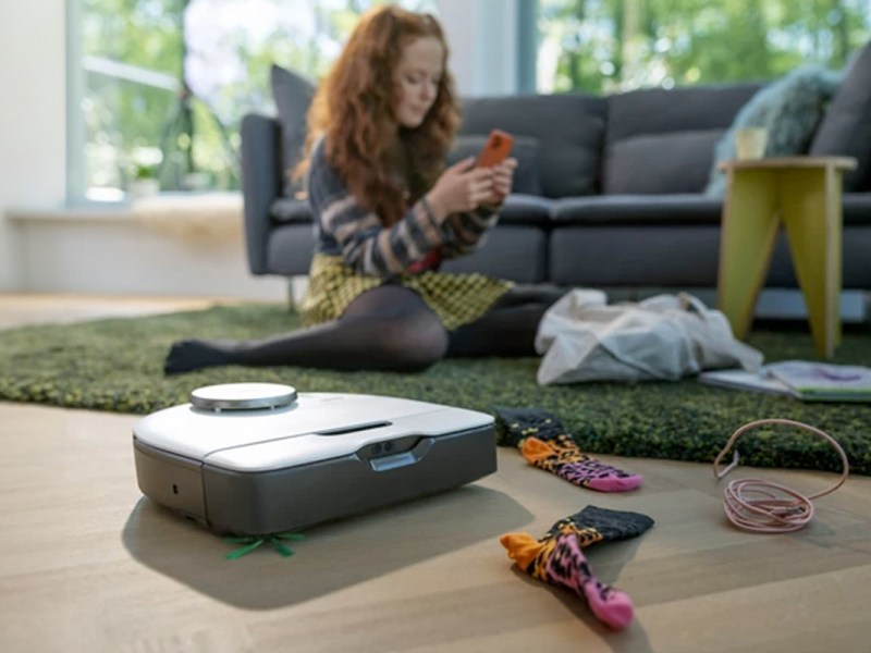 Der Vorwerk Kobold VR7 fahrt in einem Wohnzimmer. Im Hintergrund sitzt ein Mädchen auf dem Teppich und spielt. Vor dem Roboter liegen Hindernisse, wie Socken.