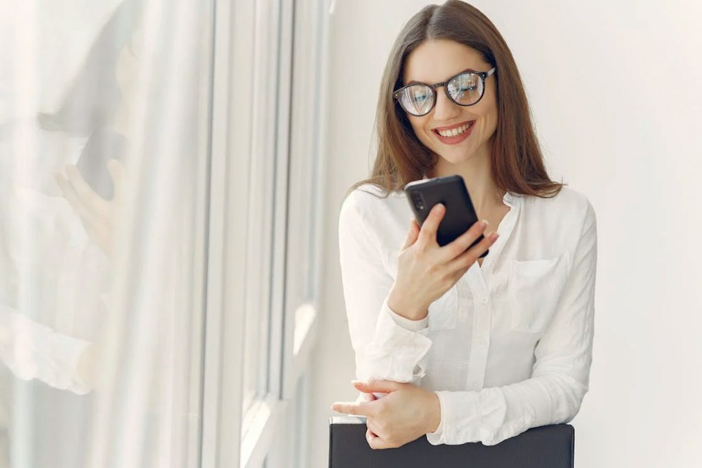 Eine Frau mit Brille steht am Fenster und schaut auf ihr Handy.