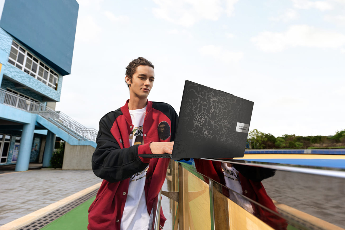 Ein sportlicher Mann guckt auf ein Notebook, das auf der Brüstung einer Sportanlage steht, im Hintergrund ein blaues Haus mit Treppenaufgang