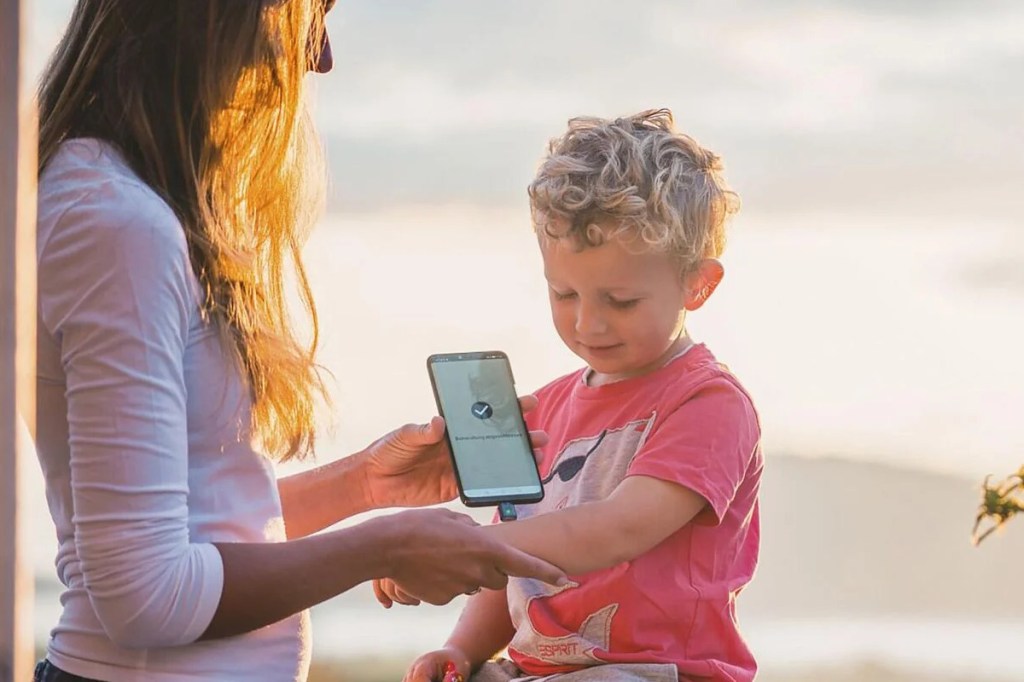 Frau steht neben sitzendem Kind und hält Smartphone mit angedocktem Heat-It-Stichheiler an Arm