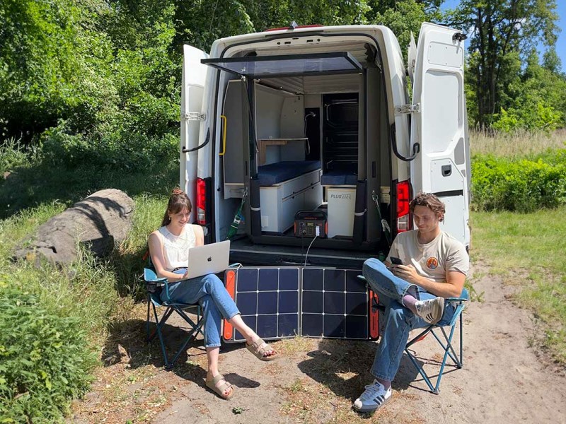 Zwei Personen sitzen vor einem Transporter, der mit einem PlugVan-Modul ausgestattet ist. Außerdem nutzen sie mobile Endgeräte, die mit einer Jackery Powerstation versorgt werden.