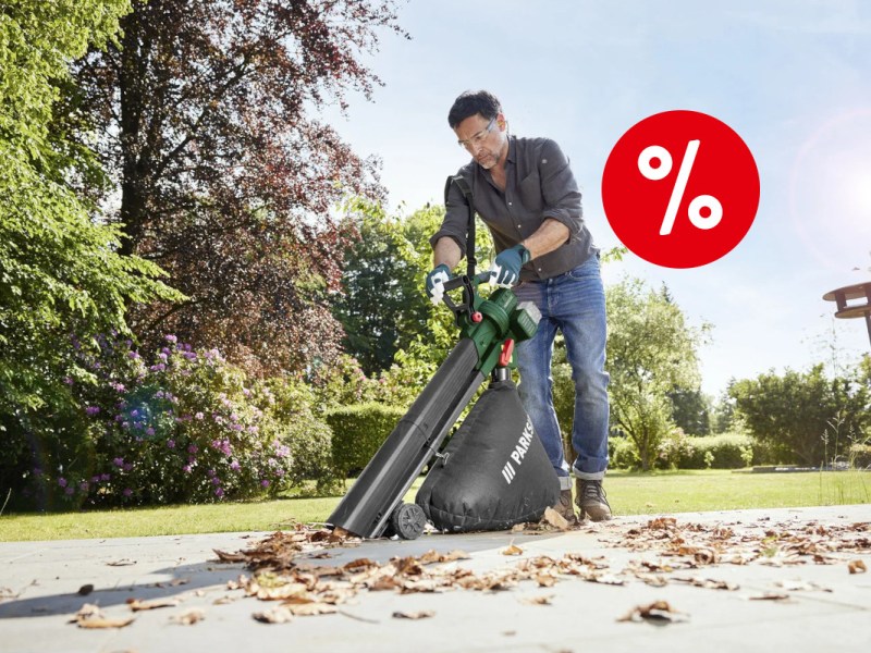 Mann mit kurzen schwarzen Haaren mittleren Alters in grauem Hemd und Jeans mit Schutzbrille und grünen Handschuhen bedient grün schwarzen Akku-Laubsauger auf hellem Steinweg mit Blättern vor grüner Wiese umringt von Bäumen