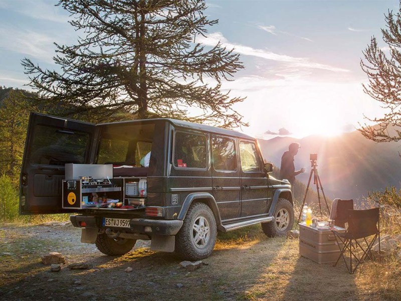 Auto steht in der Natur, Bergkulisse mit Sonnenaufgang im HIntergrund, man sieht eine Einbauküche im Heck des Autos