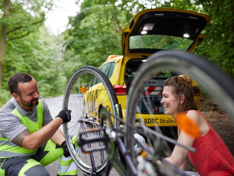 ADAC-Mitarbeiter repariert Fahrrad, lächelnde Frau und ADAC-Auto im Hintergrund