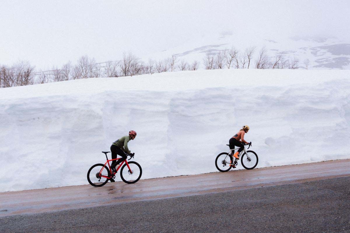 Zwei Radfahrer fahren hintereinander eine Straße lang, hinter ihnen eine Wand aus Schnee