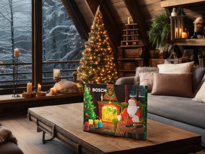 Schräg aufgestellter rechteckiger Adventskalender mit Comic-Weihnachtsmann drauf auf flachem Holztisch, der vor grauem Sofa steht, Holzboden, schräge Holzwand und Fensterfront vor winterlicher Waldkulisse im Hintergrund, daneben ein geschmückter warmleuchtender Tannenbaum