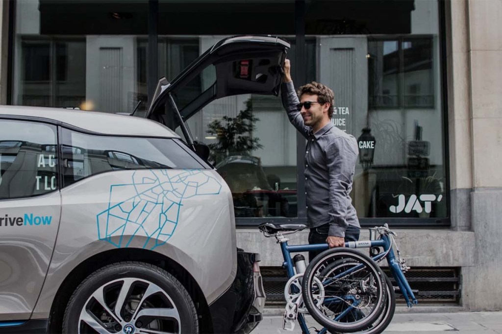 Mann steht vor einem Auto mit geöffnetem Kofferraum, in einer Hand hält er ein zusammengeklapptes Rad fest