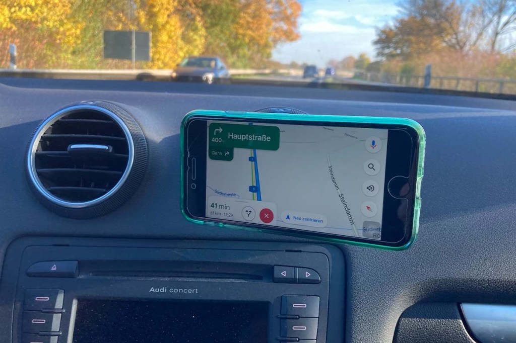 Blick aus der WIndschutzscheibe eines Autos, Smartphone mit Routenplanung im Vordergrund am Gebläse festgehalten