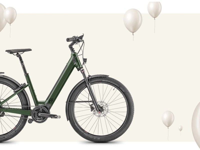 Anschnitt eines grünen E-Bikes, welches von links ins Bild gefahren kommt auf einem beigen Untergrund, dazu Luftballons