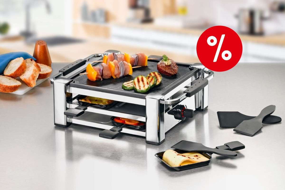 Silbern schwarzer Raclette-Grill schräg von vorne mit zwei Etagen mit befüllten Pfännchen und Gemüse oben auf einer Grillplatte auf hellem spiegelndem Tisch umringt von gebackenenm Käse in Pfännchen und Spateln