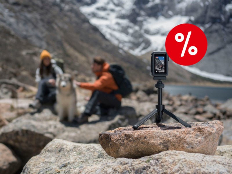 Rechts im Vordergrund eine kleine schwarze hochkant aufgestellte DJI Osmo Action 4 auf kleinem Dreibein-stativ auf Felsen platziert, im Hintergrund zwei Menschen sitzend mit Rucksäcken und weiß grauem Hund vor grau weißer Berglandschaft mit rotem Prozentbutton oben rechts