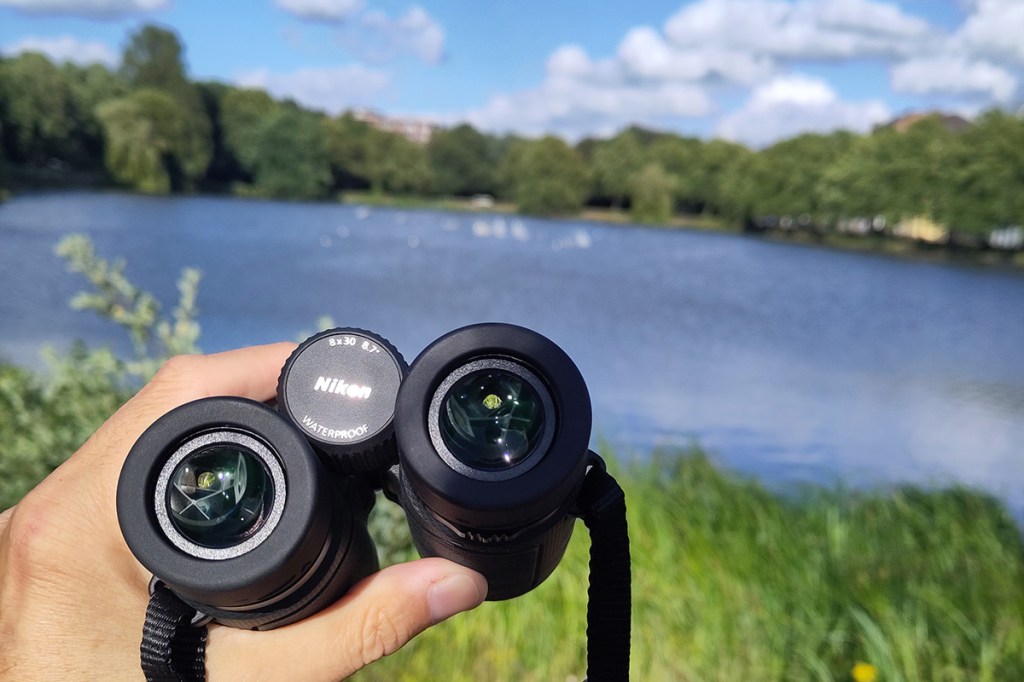 Ein Fernglas, in der linken Hand gehalten, im Hintergrund ein unscharfer blauer See.