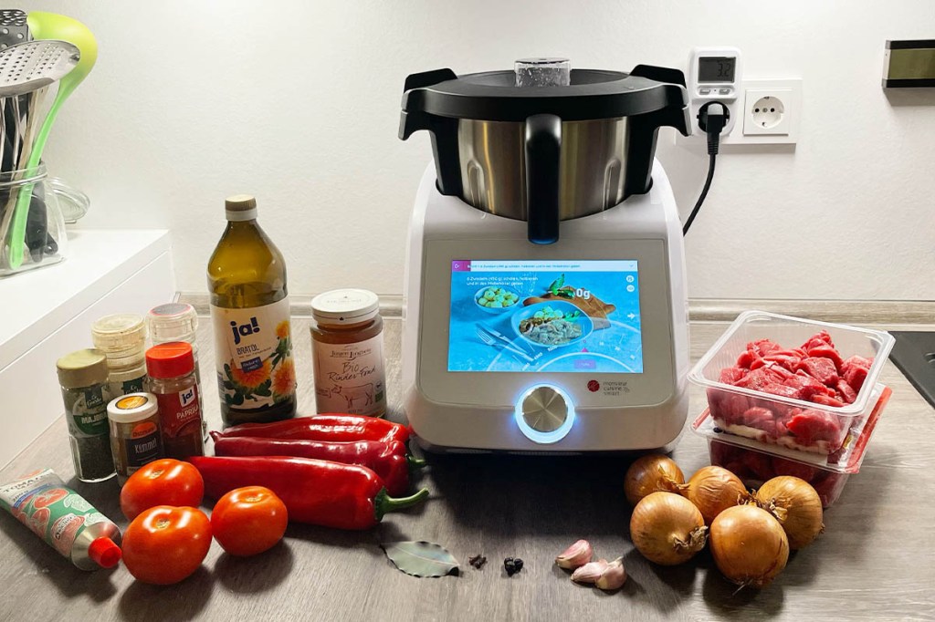 Der Monsieur Cuisine Smart mit Zutaten für ein Gulasch auf einer Arbeitsplatte in der Küche.