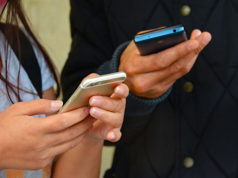 Zwei Paar Hände, links von Frau, rechts von Mann, halten jeweils ein Smartphone (gold, blau), die Frau hat ein hellblaues shirt an, der Mann eine dunkelblaue Jacke