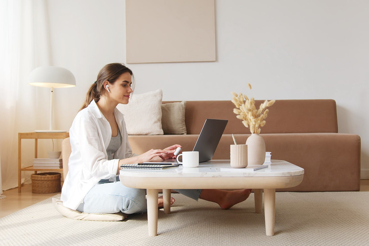 Lächelnde Frau mit Pferdeschwanz in weißem Hemd sitzt im SChneidersitz an hellem Couchtisch auf dem ein aufgeklappter Laptop und Tassen und Vasen stehen, von der Seite, dahinter eine ocker farbene Couch und helle Wand