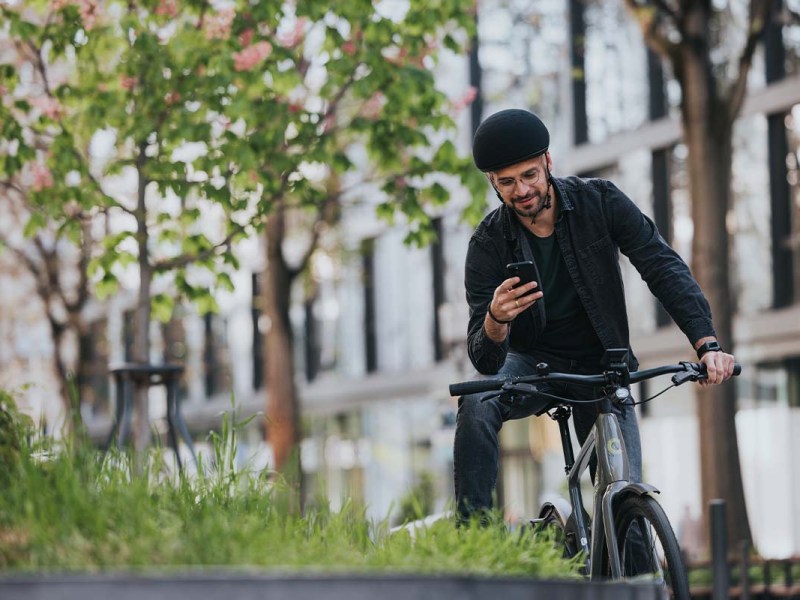 Mann sitzt auf seinem Rad, hat den einen Fuß auf einer Mauer gelehnt, er schaut in sein Smartphone