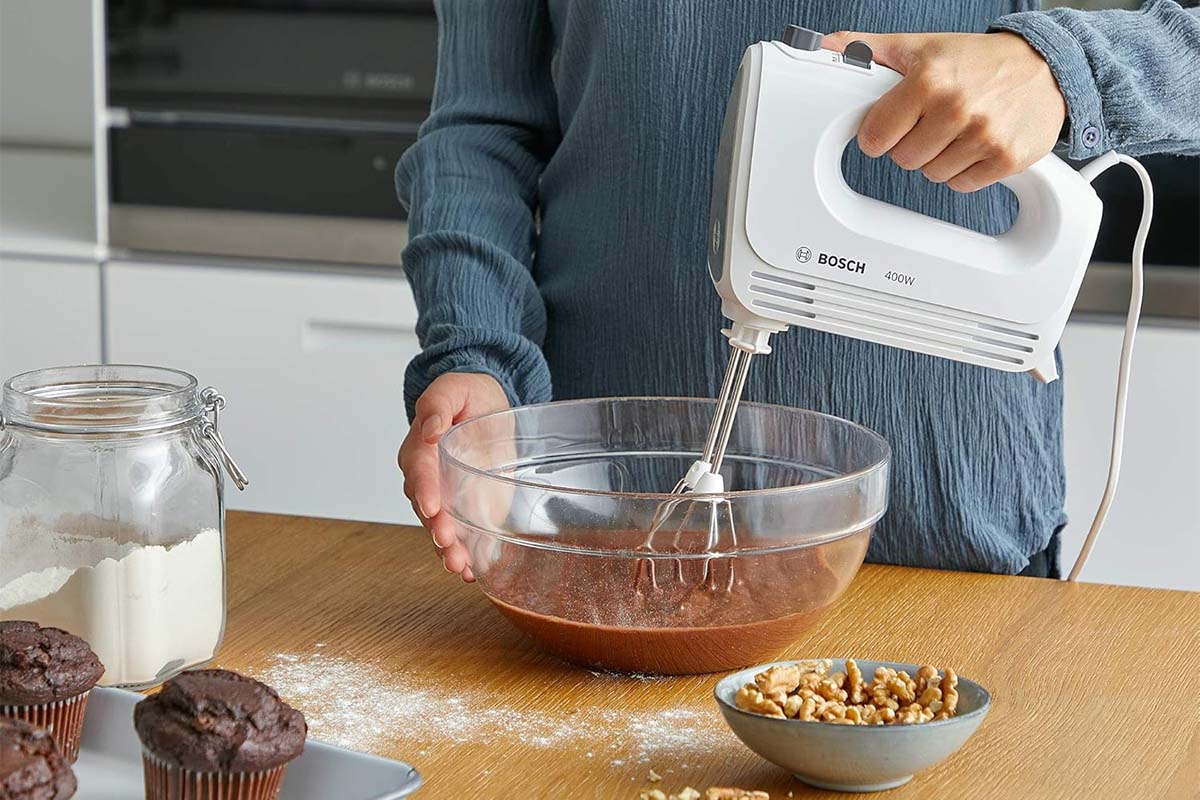 Person im Anschnitt, hat einen Handmixer in der Hand und rührt einen braunen Teig