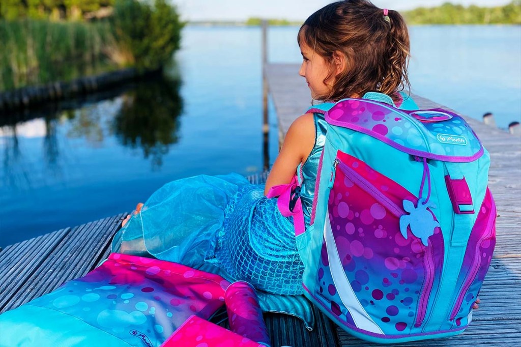 Mädchen mit Schulranzen auf dem Rücken von hinten, auf einem Steg am See sitzend